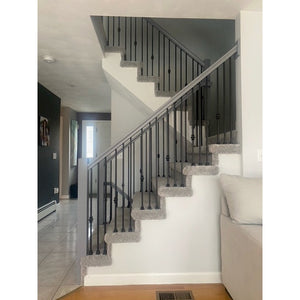 A carpeted staircase features the Holmes Contemporary 2-5/8"W x 1-3/4"H 6084 Handrail with black metal railings. The stairs turn at a landing, with light walls, a tiled floor, and part of a beige sofa nearby in this modern home.